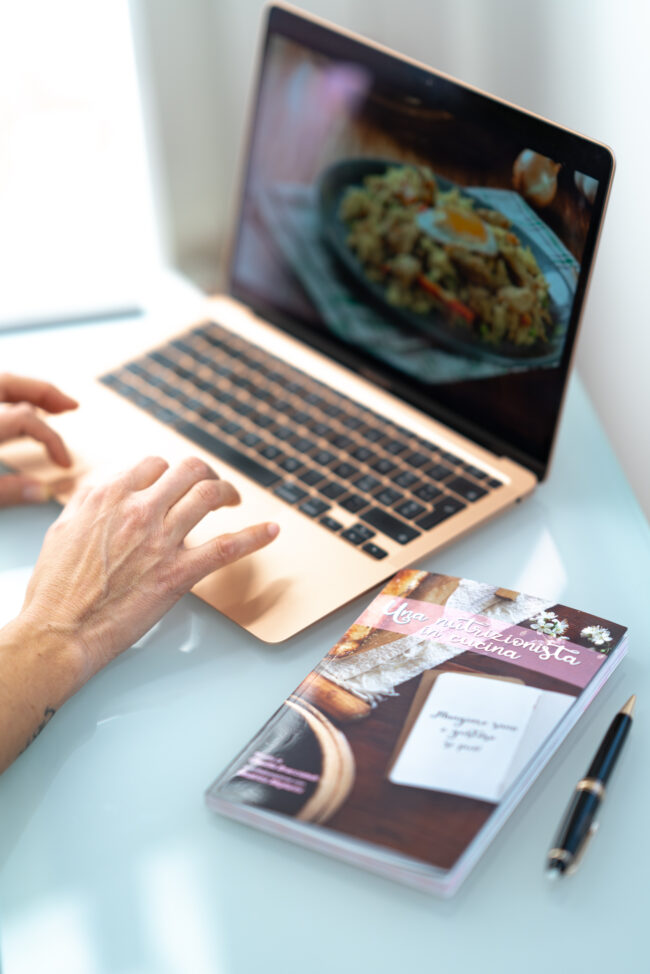 Mani di una Nutrizionista al Lavoro al Computer con un Libro di Nutrizione Affianco, Un'Immagine che Rappresenta l'Impegno e la Costante Formazione nell'Ambito della Salute Alimentare.