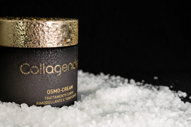 Fotografia Still Life di Crema Tonificante: Sfondo Nero, Confezione di Crema Bagnata da Gocce d'Acqua, appoggiata su sale grosso, un Dettaglio Accattivante che Evidenzia Freschezza e Benessere.