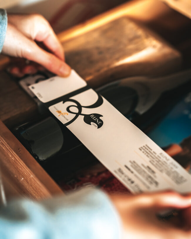 Precisione Artigianale: Mani che Attaccano un'Etichetta a una Bottiglia di Vino, un Momento Cruciale nel Processo di Imbottigliamento.