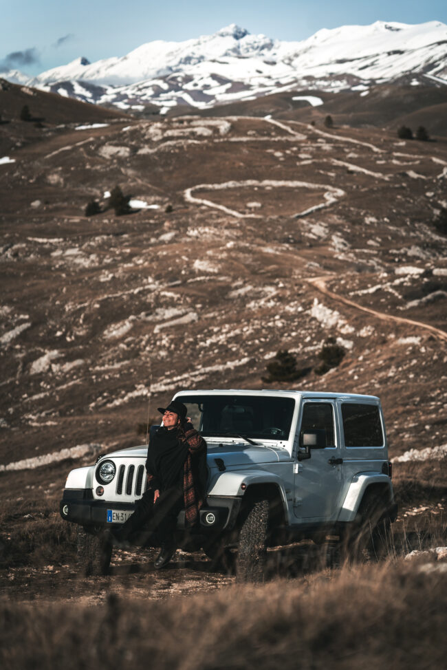 Giovane donna appoggiata su una Jeep Wrangler bianca, indossa cappello e sciarpa con il viso rivolto verso il sole, sfondo di montagne innevate.