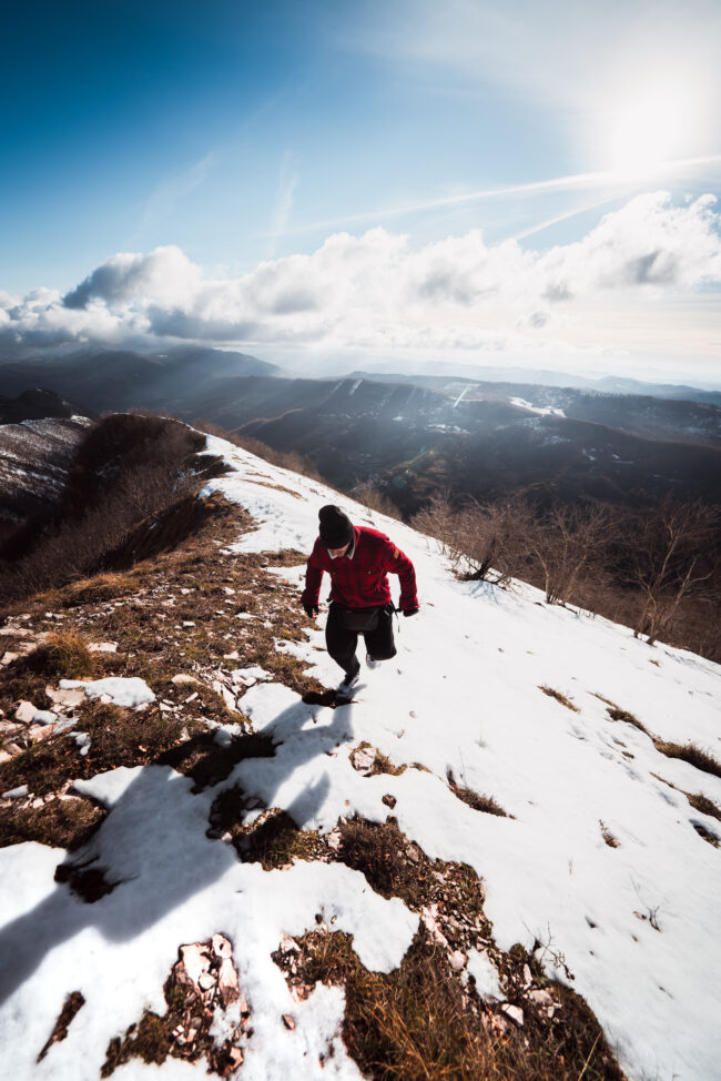 Ragazzo che cammina in salita lungo la cresta di una montagna innevata, indossando abbigliamento invernale sotto un cielo incredibile.