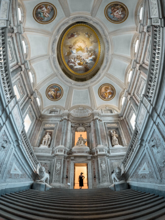 Ragazza sulle scale della sala principale della Reggia di Caserta, di fronte a una porta illuminata.